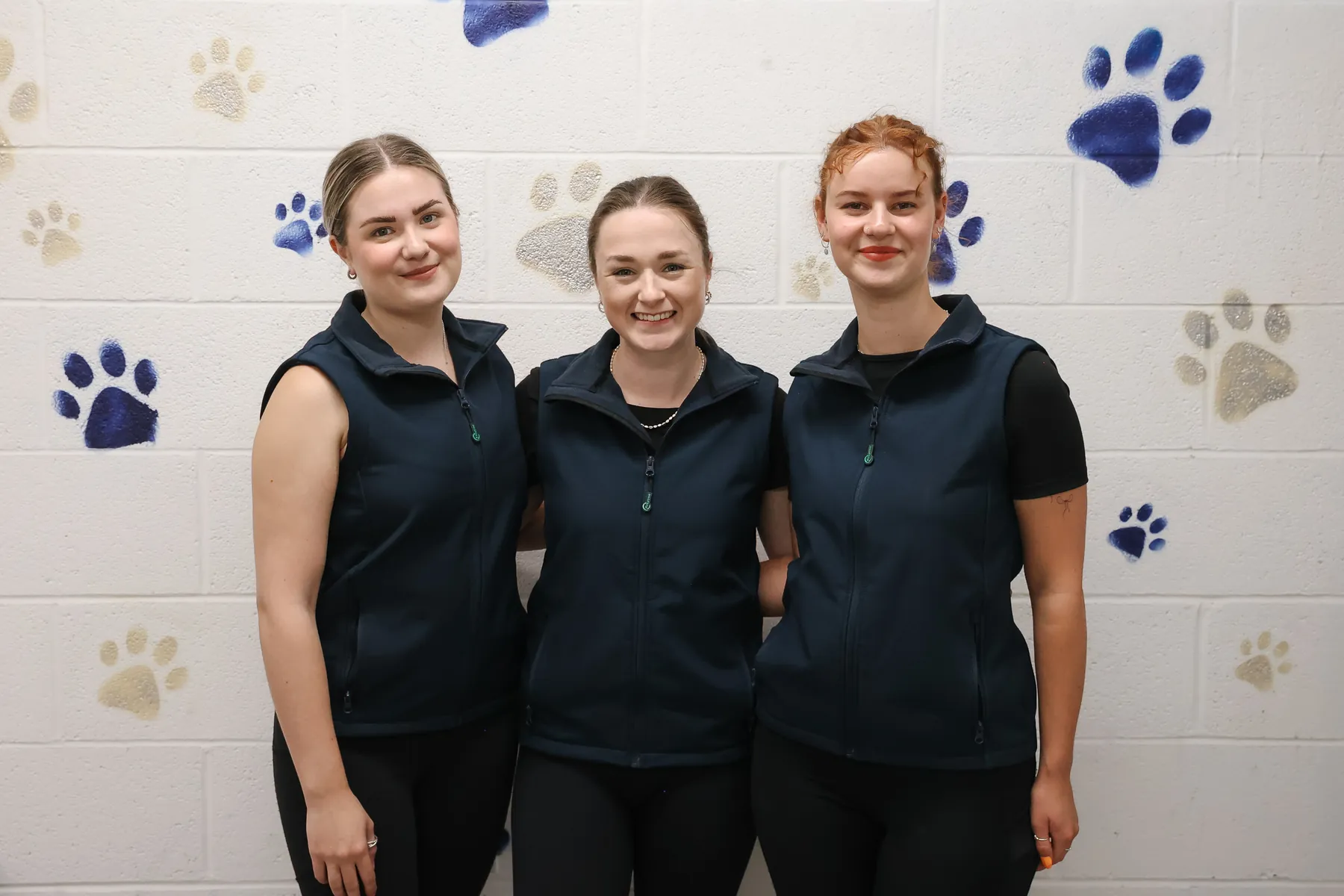 Meg, Georgie and Ruby standing together in their navy gilets in front of the paw-print wall of the groom room